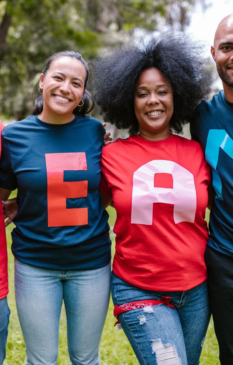 A diverse group of adults bonding and smiling in a park wearing TEAM shirts.